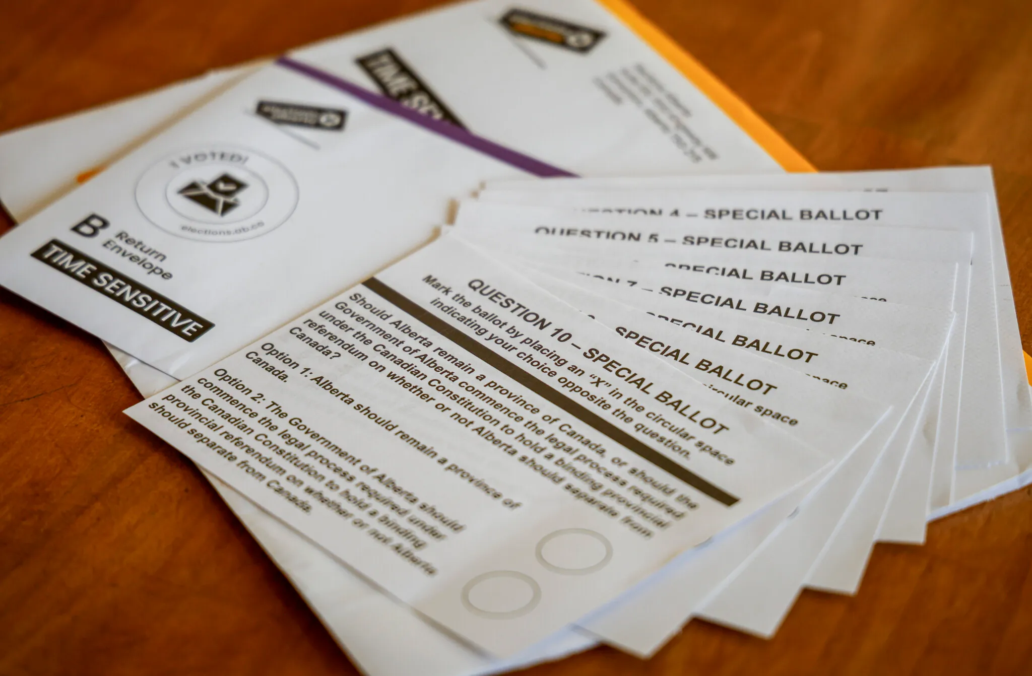 Alberta referendum special ballots for questions 4 through 10 fanned out on a wooden table beside a time-sensitive mail-in return envelope.